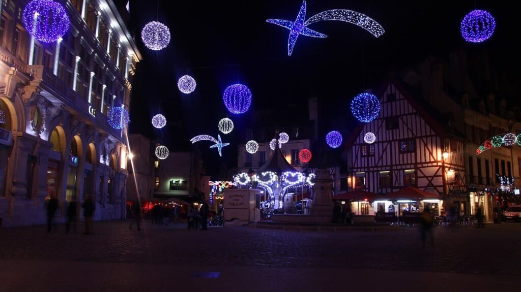 La place François-Rude illuminée pour Noël, avec ses boules et étoiles filantes lumineuses, son manège et sa maison à colombages