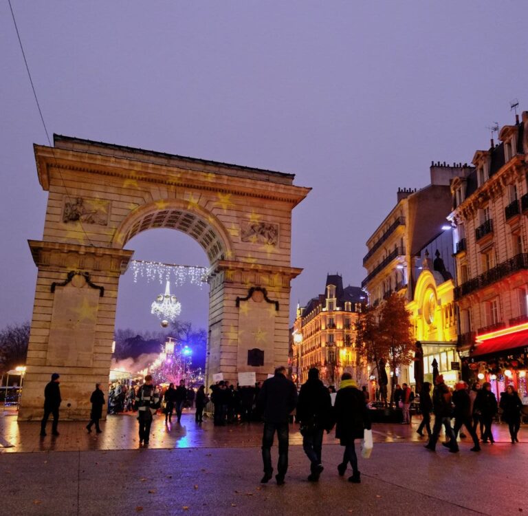 La porte Guillaume place Darcy au crépuscule, avec un lustre lumineux suspendu sous l’arche et les promeneurs de décembre