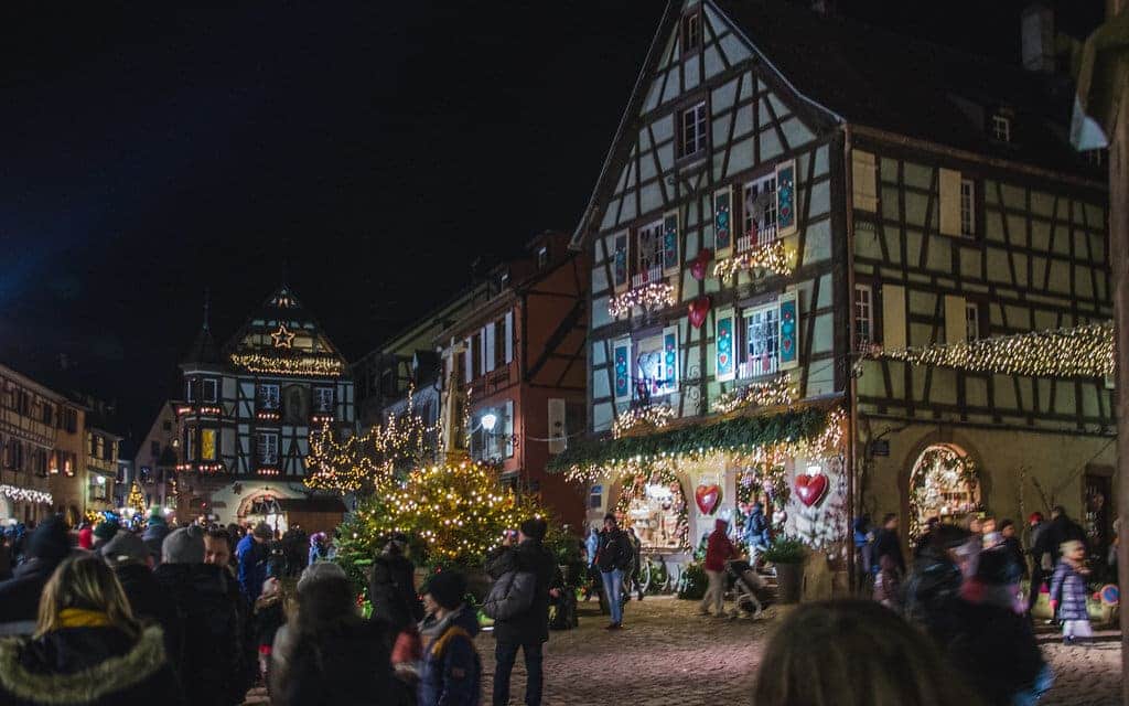 La place Jean-Ittel la nuit : maisons à colombages décorées, sapin illuminé et visiteurs du marché de Noël