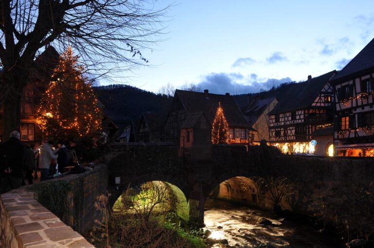 Le pont de pierre sur la Weiss, éclairé par en dessous, avec les maisons du village et un sapin lumineux au crépuscule