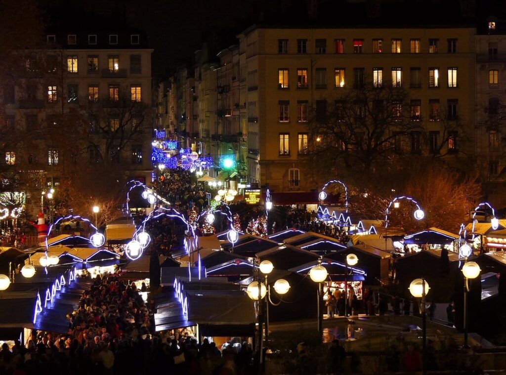 Le marché de Noël de la place Carnot vu d’en haut un soir de décembre, chalets couronnés de guirlandes bleues et immeubles lyonnais éclairés