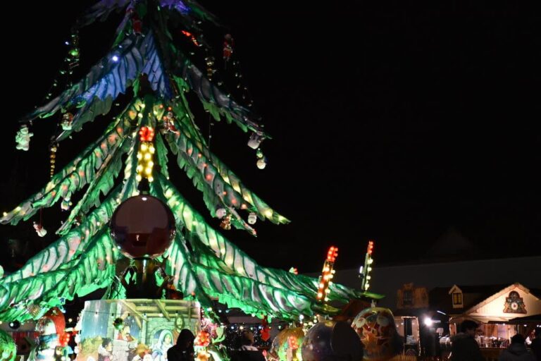 Sapin décoré de grandes feuilles lumineuses et de boules au milieu des chalets, de nuit