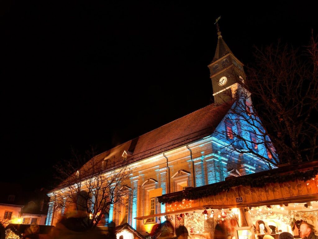 Le temple Saint-Martin de Montbéliard illuminé de bleu et de rouge, avec les chalets du marché de Noël à son pied