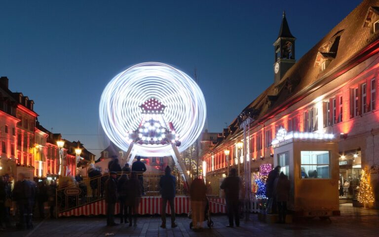 Grande roue illuminée au bout d’une place bordée de façades éclairées, à l’heure bleue