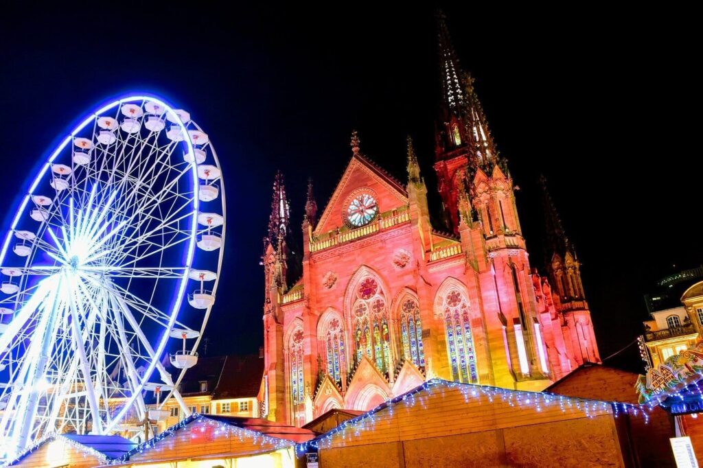Le temple Saint-Étienne illuminé en rouge et la grande roue au-dessus des chalets du marché de Noël de Mulhouse, de nuit