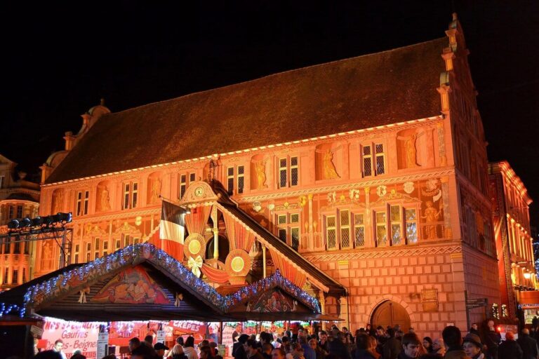 La façade peinte de l’ancien hôtel de ville de Mulhouse illuminée, derrière un chalet décoré du marché de Noël