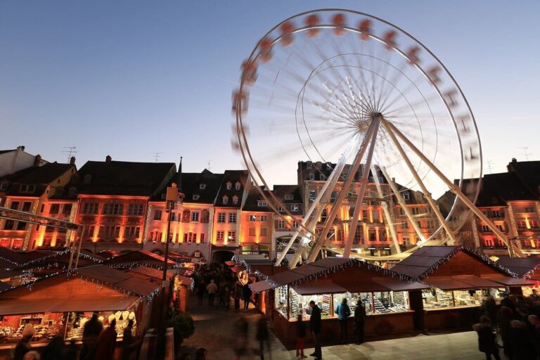 Grande roue et chalets du marché de Noël devant les façades éclairées de la place, au crépuscule