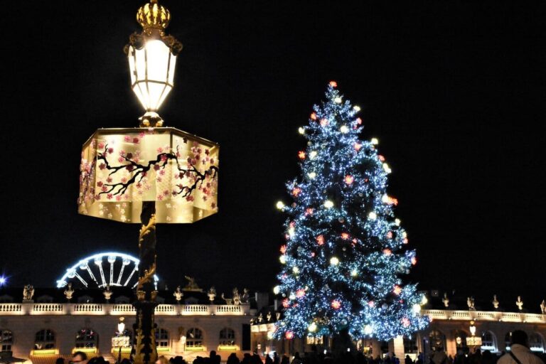 Lanterne décorée au premier plan, sapin bleu illuminé et grande roue derrière les pavillons de la place Stanislas