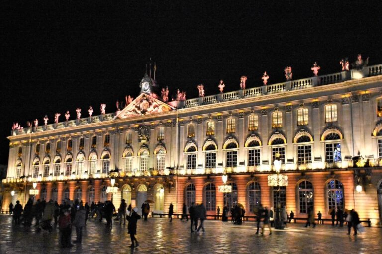 La façade de l’hôtel de ville de Nancy illuminée, avec des promeneurs sur la place Stanislas mouillée