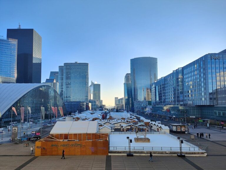 Vue d’ensemble des chalets blancs du marché de Noël sur le parvis de La Défense, entre les tours, par temps clair