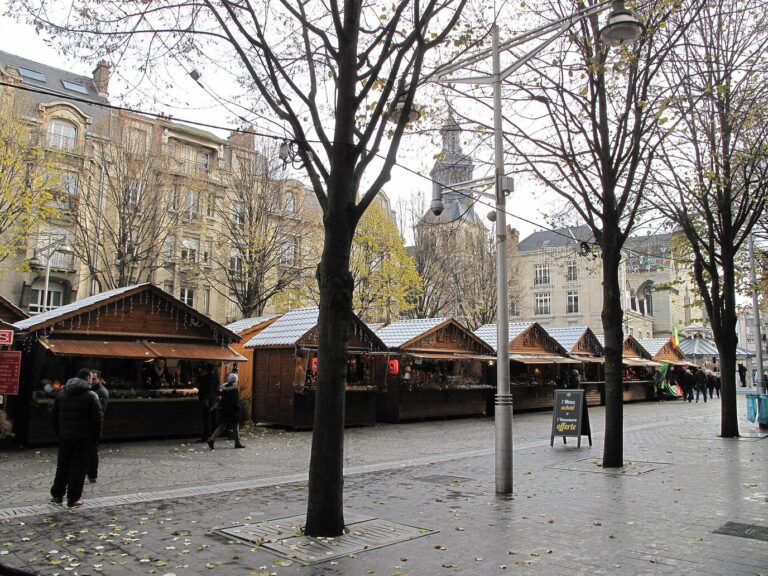 Chalets en bois du marché de Noël alignés sous les arbres de la place Drouet-d’Erlon, avec un clocher en arrière-plan