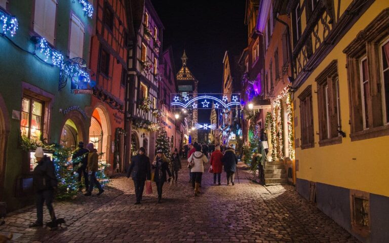 La rue du Général-de-Gaulle à Riquewihr à la tombée de la nuit, guirlandes et sapins devant les façades, avec le Dolder au fond