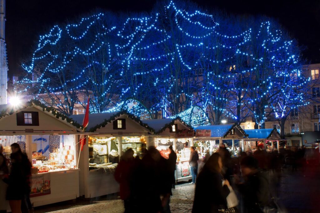 Chalets illuminés du marché de Noël de Rouen sous des arbres couverts de guirlandes bleues, le soir