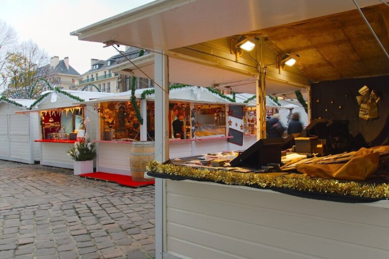 Étal d’un chalet blanc décoré de guirlandes dorées, avec d’autres chalets du marché de Noël sur la place pavée