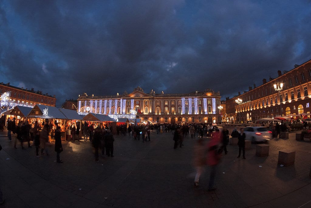 Vue panoramique de la place du Capitole sous un ciel d’orage à la tombée du jour, avec les chalets du marché et le Capitole illuminé au fond