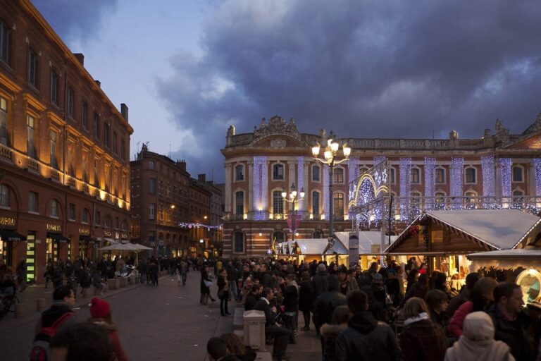 La place du Capitole au crépuscule, avec la façade du Capitole illuminée, les chalets du marché de Noël et la foule des promeneurs