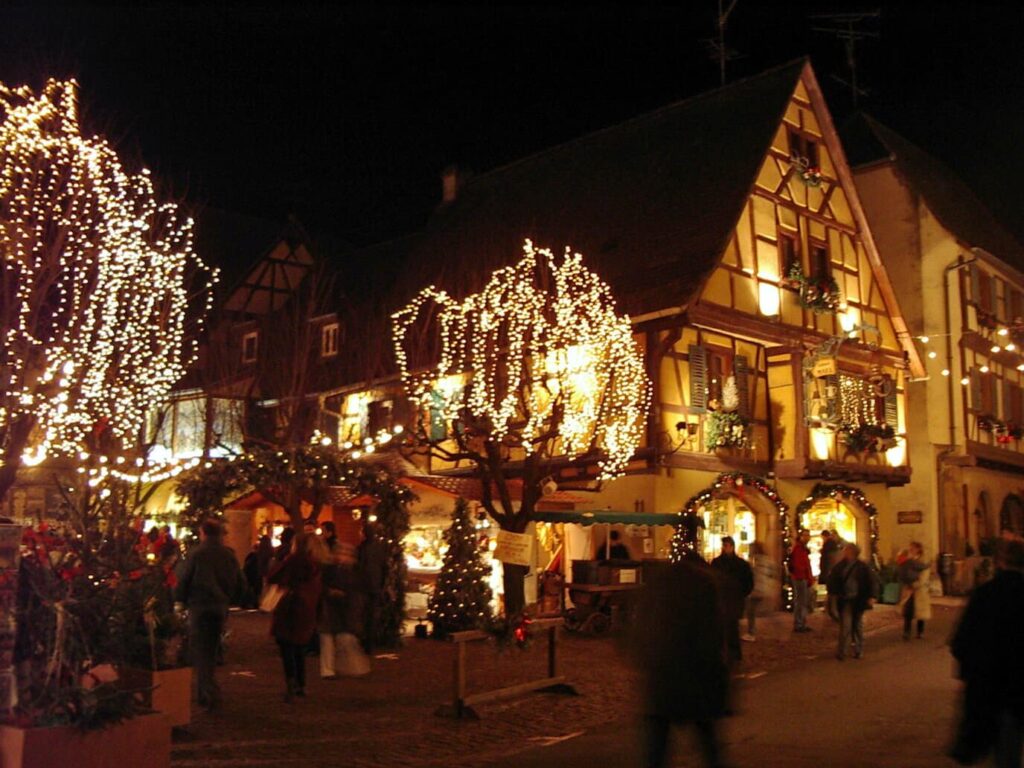Maisons à colombages d’Eguisheim illuminées de nuit, arbres habillés de guirlandes et promeneurs sur la place pendant le marché de Noël