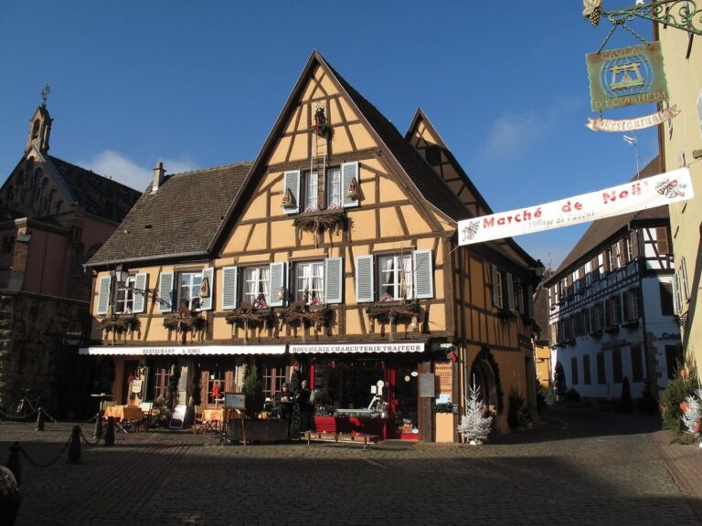 Grande maison à colombages ocre d’Eguisheim sous un ciel bleu, avec une banderole « Marché de Noël » tendue au-dessus de la rue