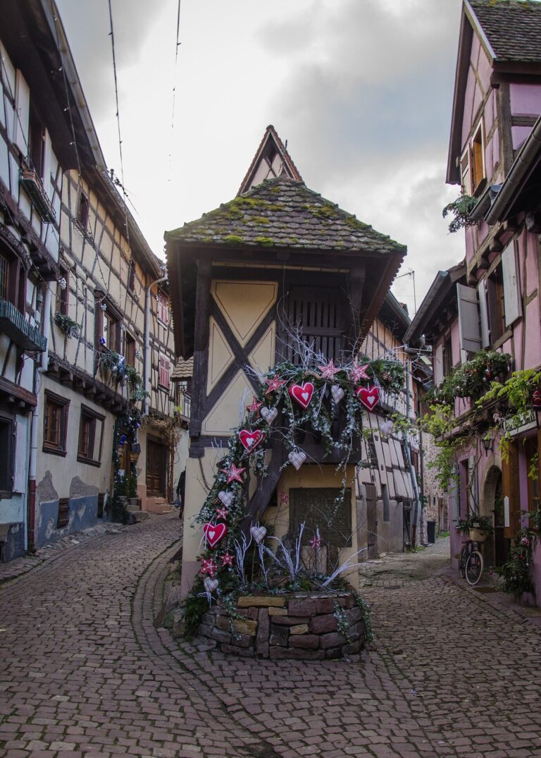 Petite maison à colombages au croisement de deux ruelles pavées, avec une fontaine décorée de cœurs pour Noël