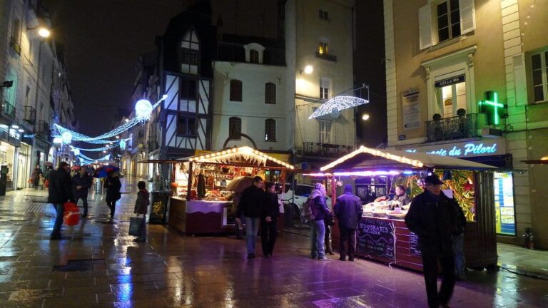 Chalets éclairés du marché de Noël sur la place du Pilori, devant des maisons à pans de bois, un soir de pluie
