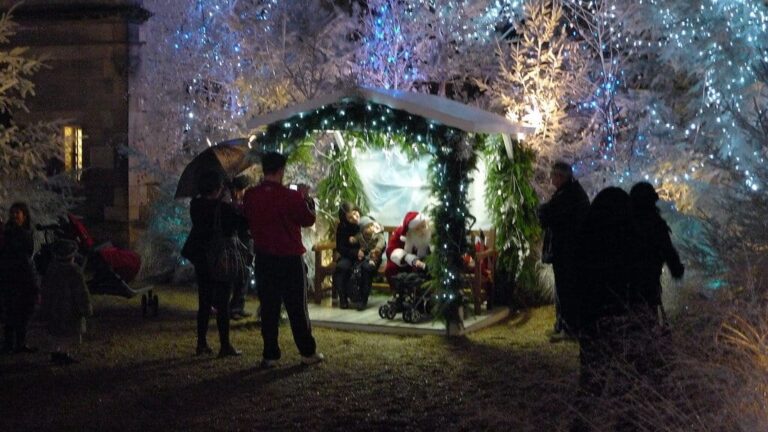 La cabane du Père Noël entourée d’arbres givrés et lumineux, avec des familles venues le rencontrer, de nuit