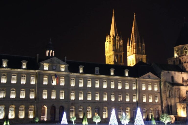 La façade éclairée de l’hôtel de ville et les flèches de l’abbatiale Saint-Étienne, devant des sapins lumineux
