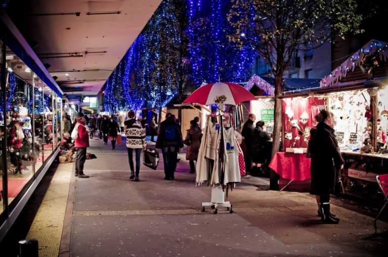 Allée de chalets décorés le long d’une galerie commerçante, sous des arbres couverts de guirlandes bleues