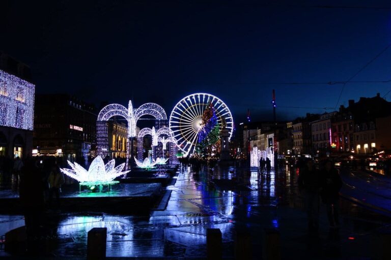 Décors lumineux en forme de fleurs et d’arches et grande roue sur la place de Jaude, reflétés sur le sol mouillé