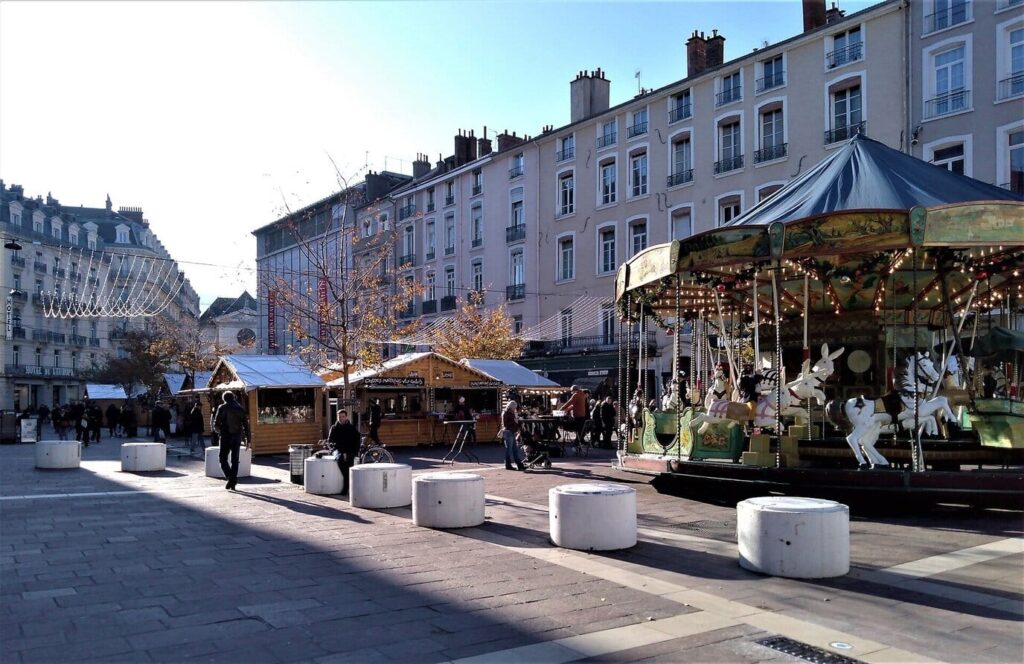 Le manège ancien et les chalets du marché de Noël sur la place Grenette, sous un ciel d’hiver dégagé, avec les guirlandes tendues au-dessus de la rue