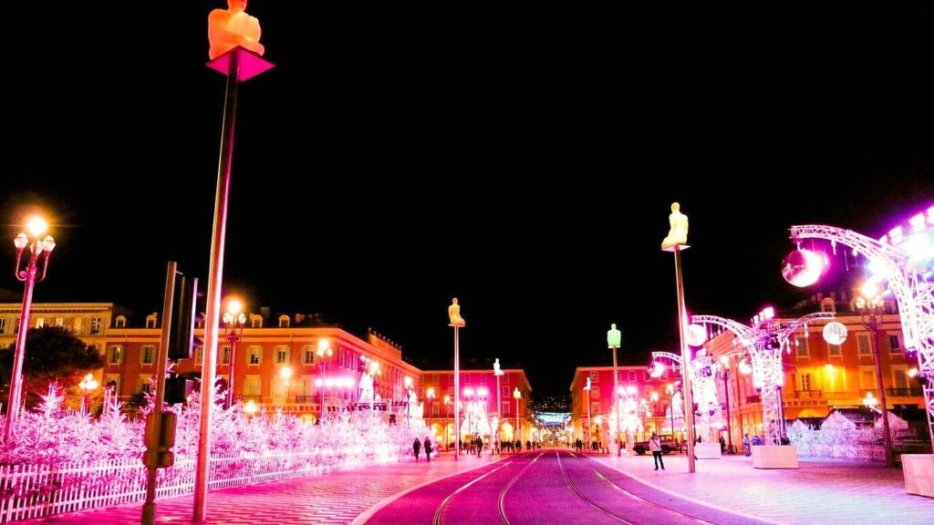 La place Masséna illuminée pour Noël, ses façades rouges, ses statues lumineuses perchées sur leurs mâts et ses sapins blancs éclairés de rose