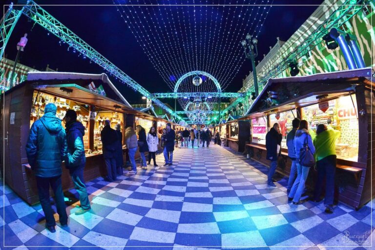 Une allée de chalets du village de Noël sur un sol en damier, sous des arches et des guirlandes lumineuses, place Masséna