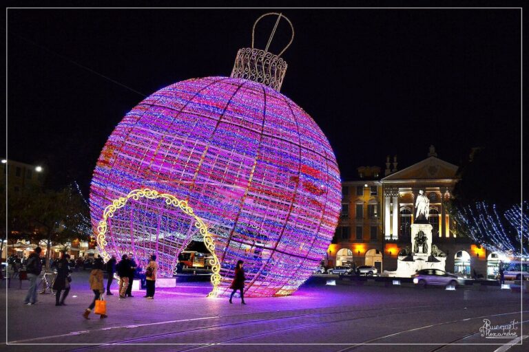 Une boule de Noël géante en lumières violettes et roses, que les promeneurs traversent par une arche, devant un monument éclairé