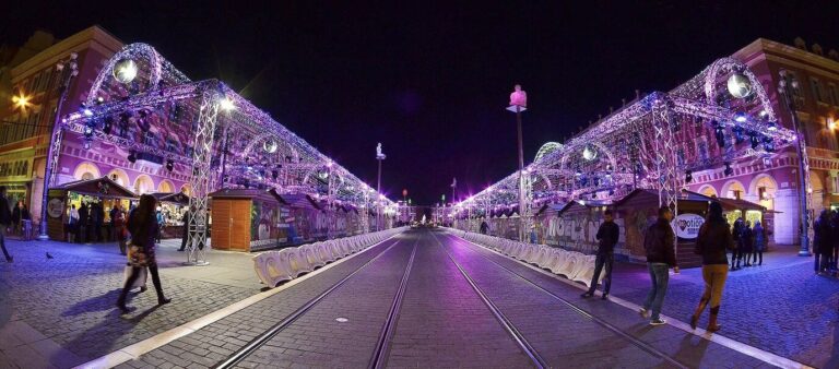 Deux rangées de chalets sous des arches lumineuses violettes, de part et d’autre des rails du tramway