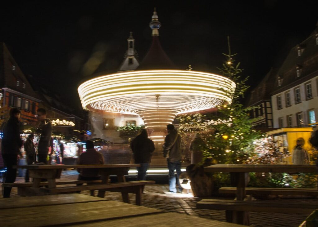 Le carrousel illuminé tourne au milieu des chalets et des tables de la place du Marché d’Obernai, à côté d’un sapin décoré, de nuit