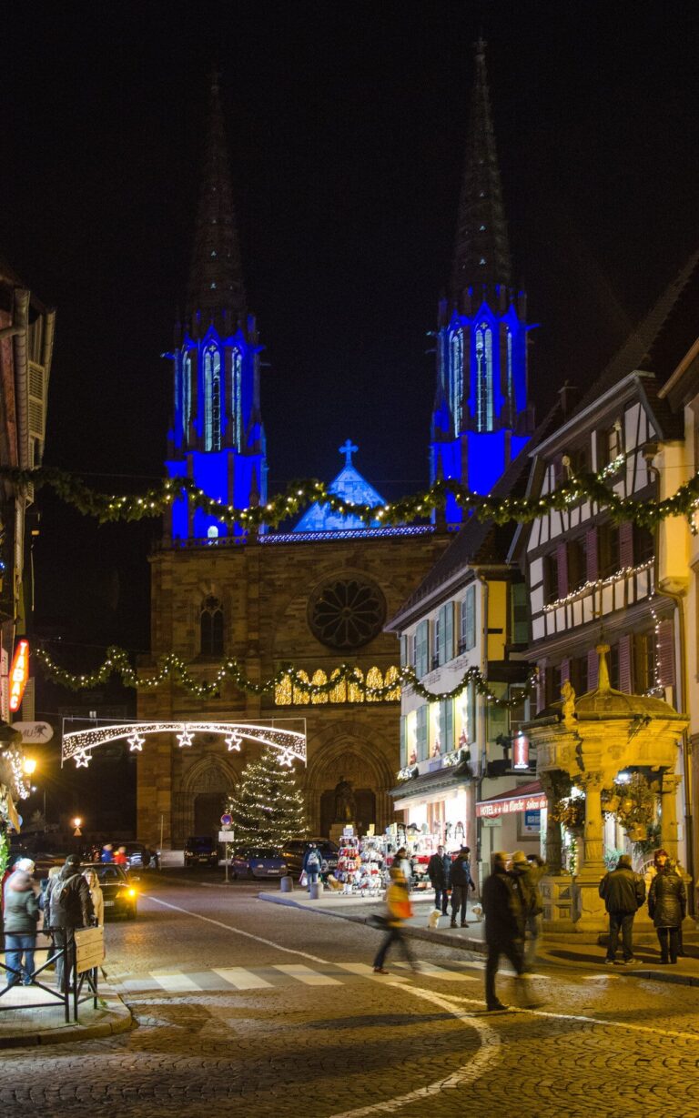 Les flèches de l’église Saints-Pierre-et-Paul éclairées en bleu au bout d’une rue décorée de guirlandes et d’un sapin lumineux