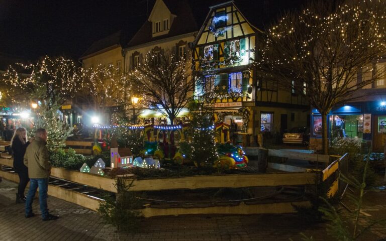 Petit train de Noël dans un décor de sapins et de maisonnettes lumineuses, devant une maison à colombages décorée