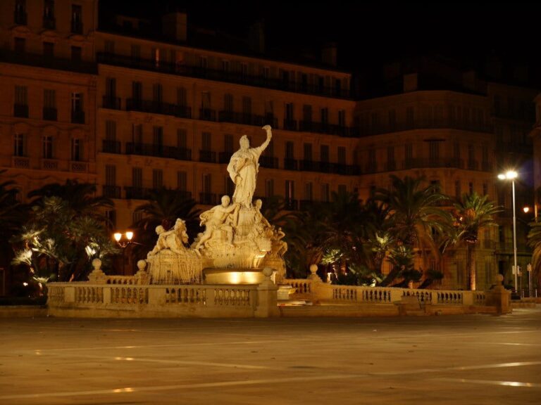 La fontaine de la Fédération éclairée la nuit sur la place de la Liberté, devant les palmiers et les immeubles de la place