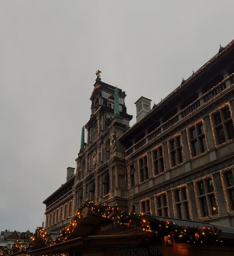 Façade de l’hôtel de ville d’Anvers soulignée de guirlandes lumineuses, au-dessus des toits décorés des chalets du marché