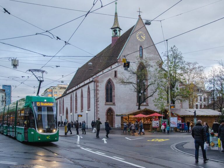 Chalets de Noël au pied de l’église Sainte-Claire (Clarakirche) de Bâle, avec un tram vert sur la Claraplatz