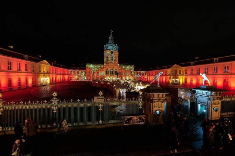 La façade du château de Charlottenburg illuminée en rouge derrière les grilles, au-dessus du marché de Noël, la nuit