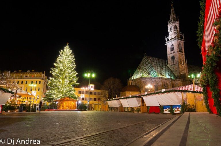 La piazza Walther de Bolzano la nuit, avec son grand sapin illuminé, les chalets du marché de Noël et le clocher de la cathédrale au toit de tuiles vernissées