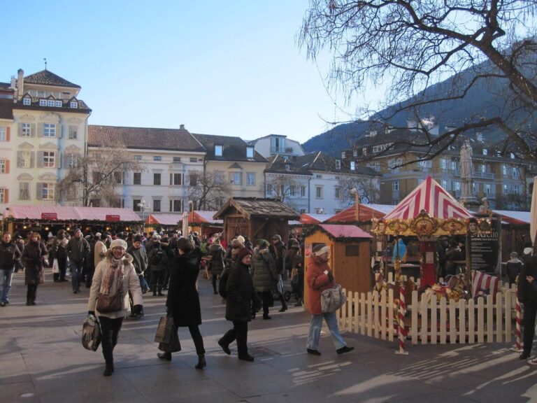 Promeneurs entre les chalets en bois et un petit manège du marché de Noël de Bolzano, sous un ciel bleu d’hiver