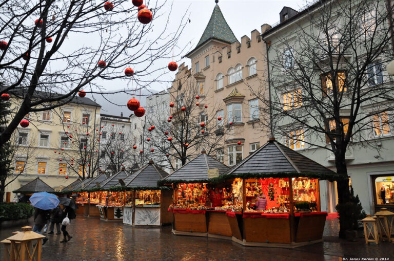 Chalets éclairés du marché sous des arbres décorés de boules rouges, devant les façades de la place