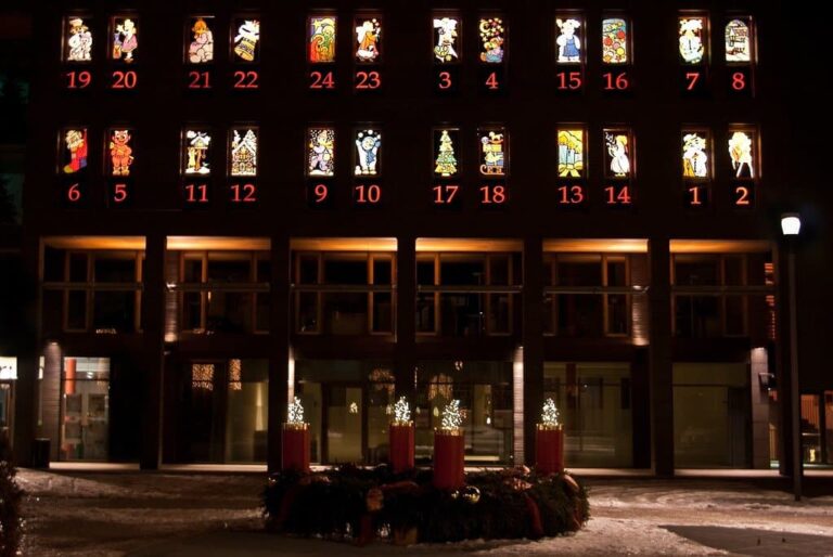 Façade transformée en calendrier de l’Avent, ses fenêtres numérotées éclairées, au-dessus d’une couronne de l’Avent à quatre bougies