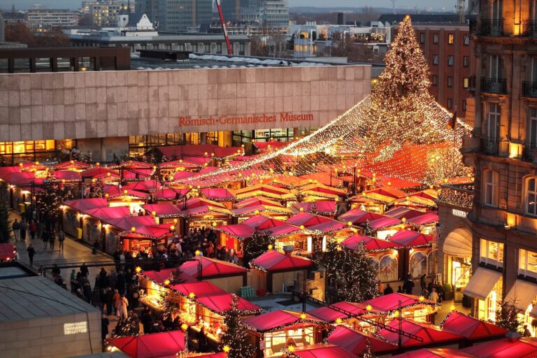 Les toits rouges des chalets du marché de la cathédrale vus d’en haut, sous une guirlande lumineuse tendue jusqu’au sapin, devant le musée romain-germanique
