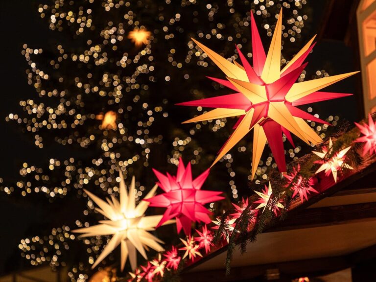 Des étoiles de Herrnhut rouges et blanches accrochées au toit d’un chalet, devant le grand sapin illuminé