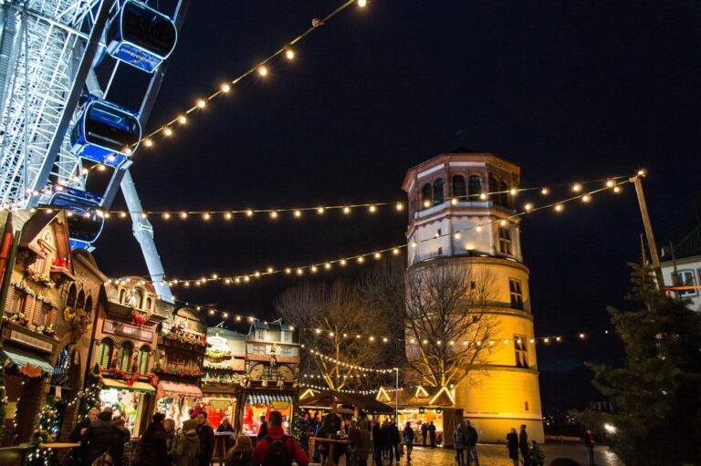 Des guirlandes d’ampoules tendues au-dessus des chalets du Burgplatz, entre la grande roue et le Schlossturm éclairé