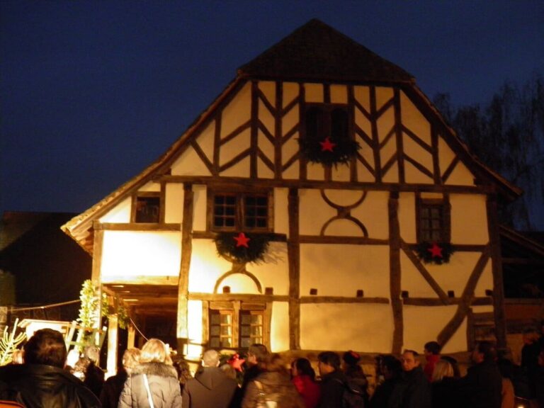 Façade à colombages ornée de couronnes de sapin et d’étoiles rouges, éclairée de nuit, devant un groupe de visiteurs vus de dos