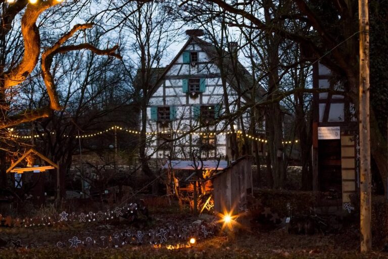 Guirlandes lumineuses et bonshommes en pain d’épices lumineux devant une maison à colombages aux volets verts, à la tombée de la nuit
