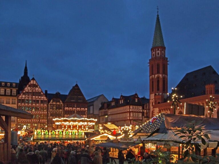 Le marché de Noël du Römerberg à l’heure bleue : manège, chalets éclairés, maisons à colombages et clocher de l’église Saint-Nicolas